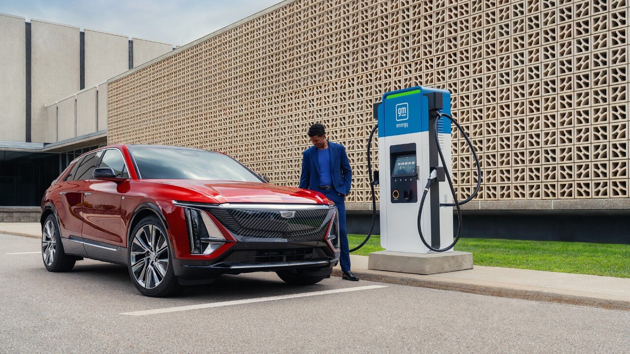 A man in a blue suit standing by a red Cadillac LYRIQ electric SUV parked at a GM EVgo charging station.