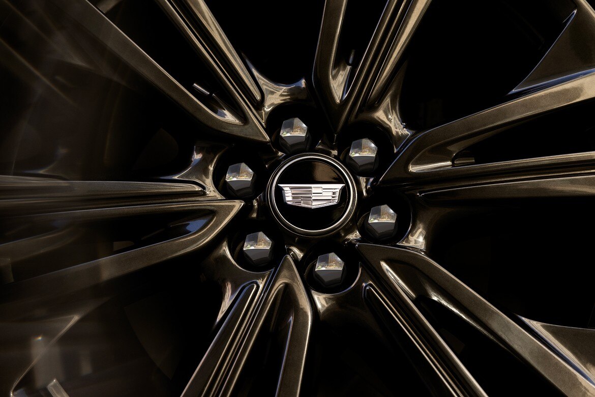 Close-up of a glossy black alloy wheel with the Cadillac emblem centered among polished lug nuts, reflecting the surrounding.